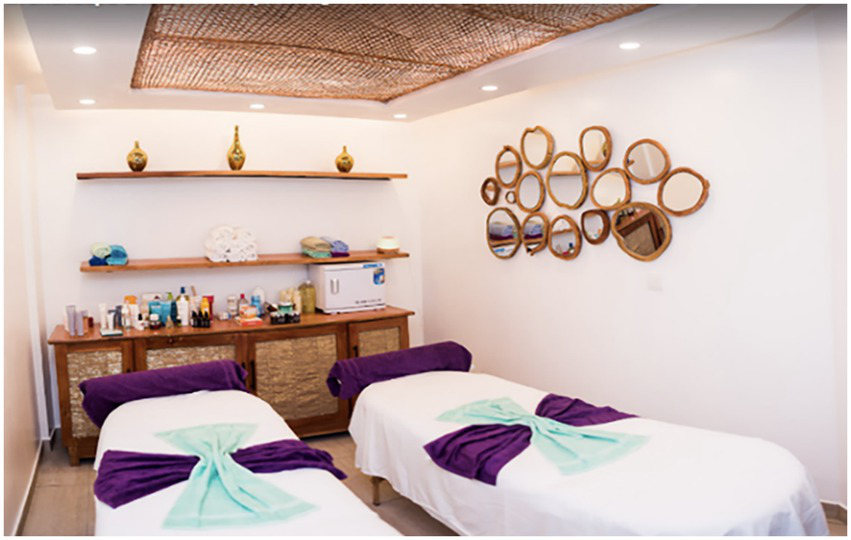 A tranquil spa room featuring two massage tables with white linens, accented by purple and mint green towels. The wall displays an arrangement of decorative round mirrors. A cabinet holds various spa products, with shelves above displaying rolled towels and small vases. The ceiling has a natural woven design, enhancing the calming atmosphere.