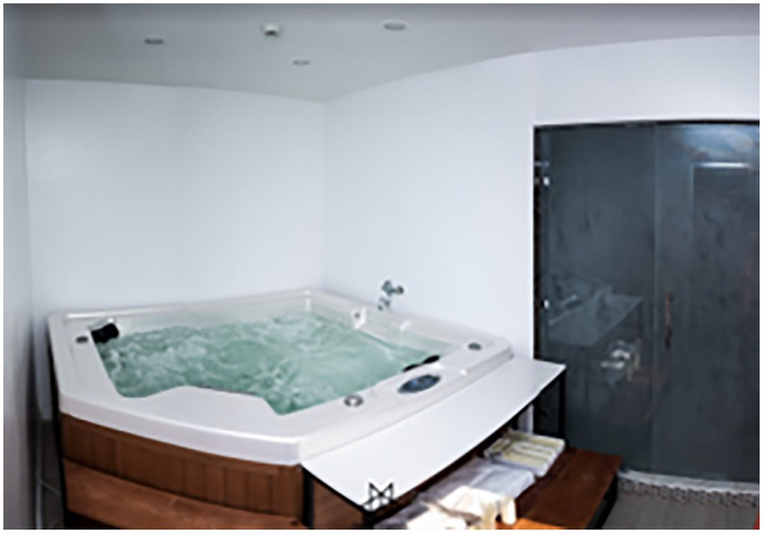 Indoor spa room featuring a large jacuzzi with bubbling water, set against white walls. A glass shower enclosure is visible to the right, alongside neatly folded towels on a wooden bench.