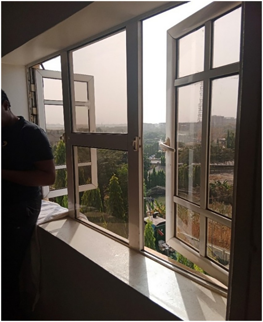 Open window with sunlight streaming in, overlooking a view of trees and distant buildings. A person stands partially visible on the left side, inside the room.