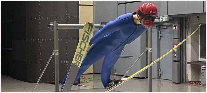 A mannequin dressed in a blue ski suit and red helmet simulates a ski-jumping pose. It is attached to a structure with support wires, with skis angled downward. The background features industrial equipment and metal doors.