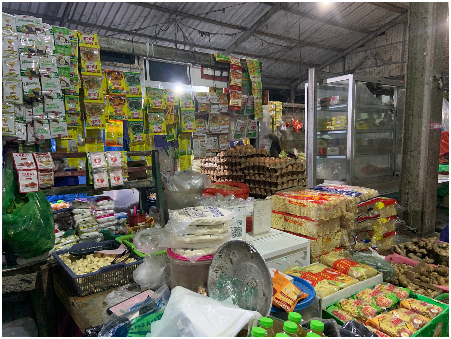 A bustling indoor market stall displaying a variety of goods. Numerous packaged snacks and spices hang on the wall. Eggs are stacked in cartons, and various dry goods and snacks are neatly arranged on the counter. A refrigerated display case is visible in the background, adding to the busy atmosphere.