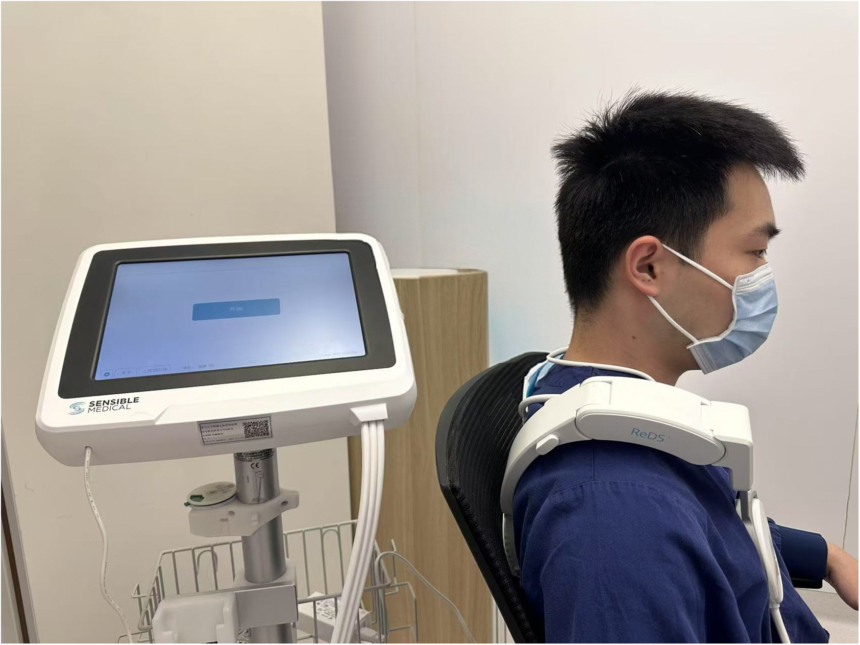 A person wearing a face mask sits in a chair with a ReDS medical device on their shoulders. A Sensible Medical monitor on a stand is nearby, displaying an interface.