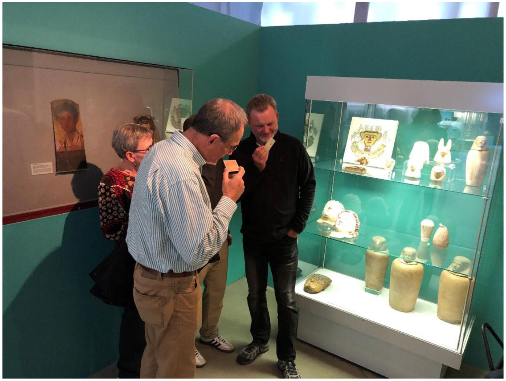 A group of people examines a museum display case containing ancient artifacts sniffing Scent of the Afterlife scent-printed cards. The case is well-lit, showcasing several items, including canopic jars. A framed painting is visible on the wall to the left. The atmosphere is focused and inquisitive.